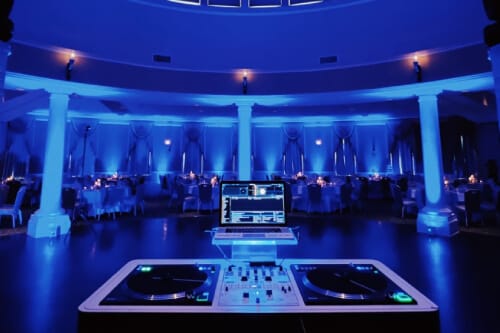 KE-Lighting-and-Design-Gallery-9-min1 A DJ booth with turntables and a laptop is set up in a dimly lit event hall with blue lighting and round tables arranged for guests.