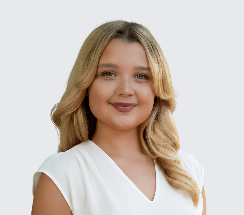 A young woman with long blonde hair and a white blouse is smiling at the camera against a plain light background.