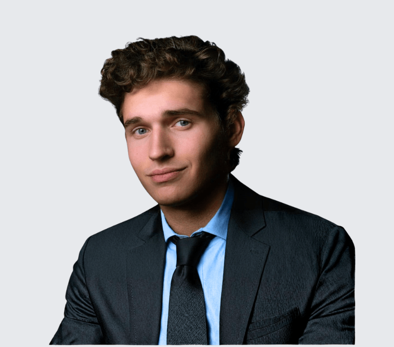 A young man with curly brown hair wearing a dark suit, light blue shirt, and black tie, posing against a plain light grey background.