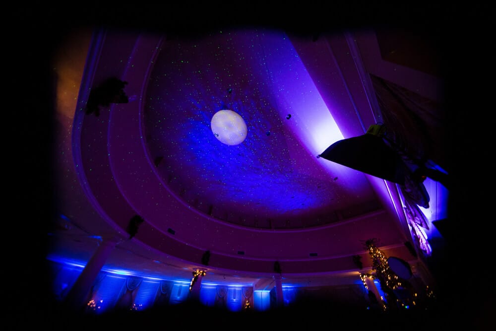 Dancing+Under+the+stars+option A domed ceiling lit with blue and purple lights, featuring a central round light fixture and star-like projections. Decorative columns and wall details are visible along the edges.