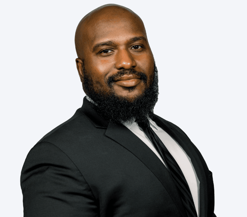 Dephilip-Jackson-Klock-Entertainment A man with a bald head and full beard wearing a black suit, white shirt, and black tie, posing against a plain light background.