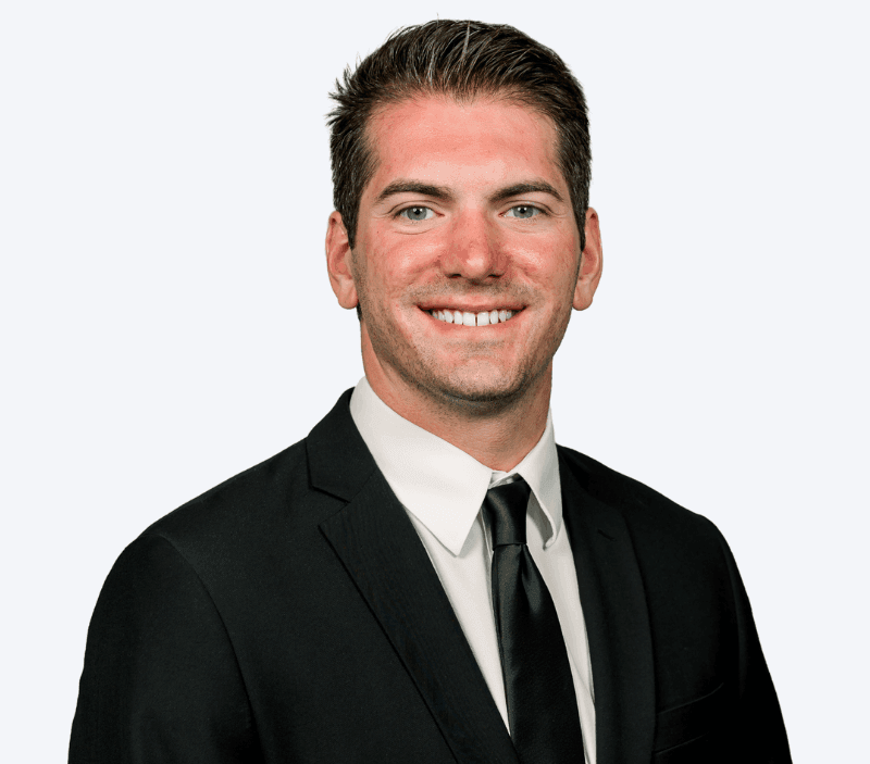 Doug-Edgar-Klock-Entertainment A man in a black suit, white shirt, and black tie is smiling at the camera against a plain light background.