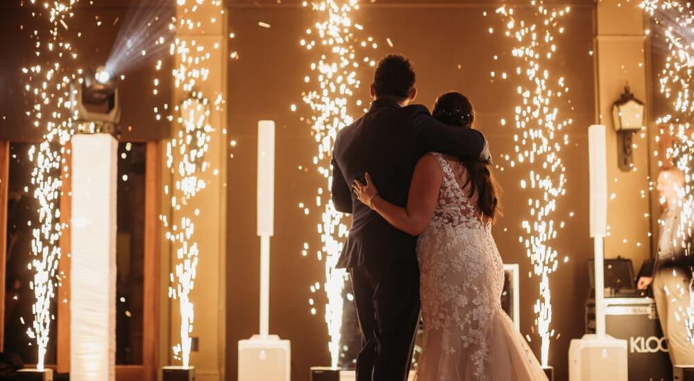 A bride and groom embrace on the dance floor surrounded by indoor fireworks during their wedding celebration.