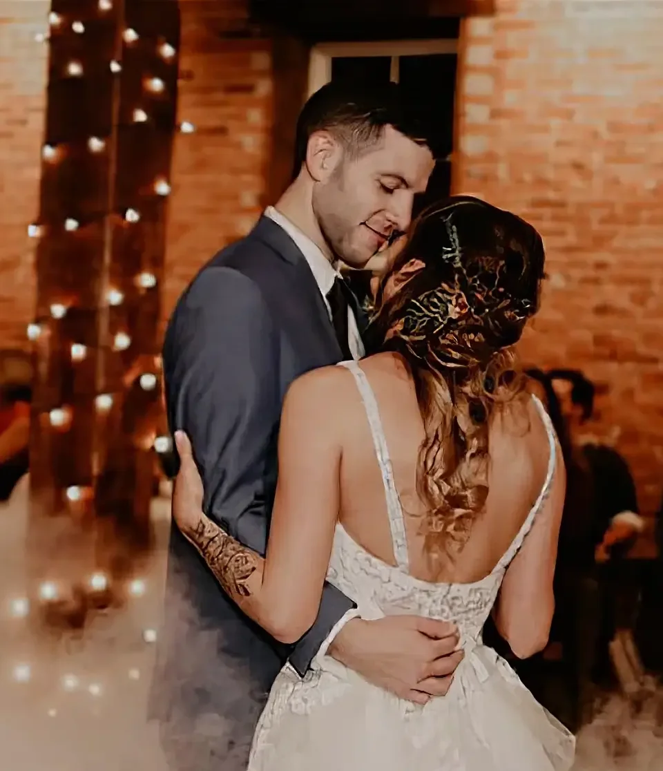 Scroll-Group-1355 A bride and groom share a dance at their wedding reception, with brick walls and string lights in the background.
