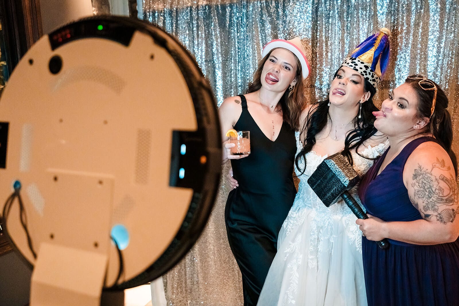 Marissa & Cory Three women, including one in a wedding dress, pose for a photo booth, wearing festive hats and holding props in front of a silver sequin backdrop.