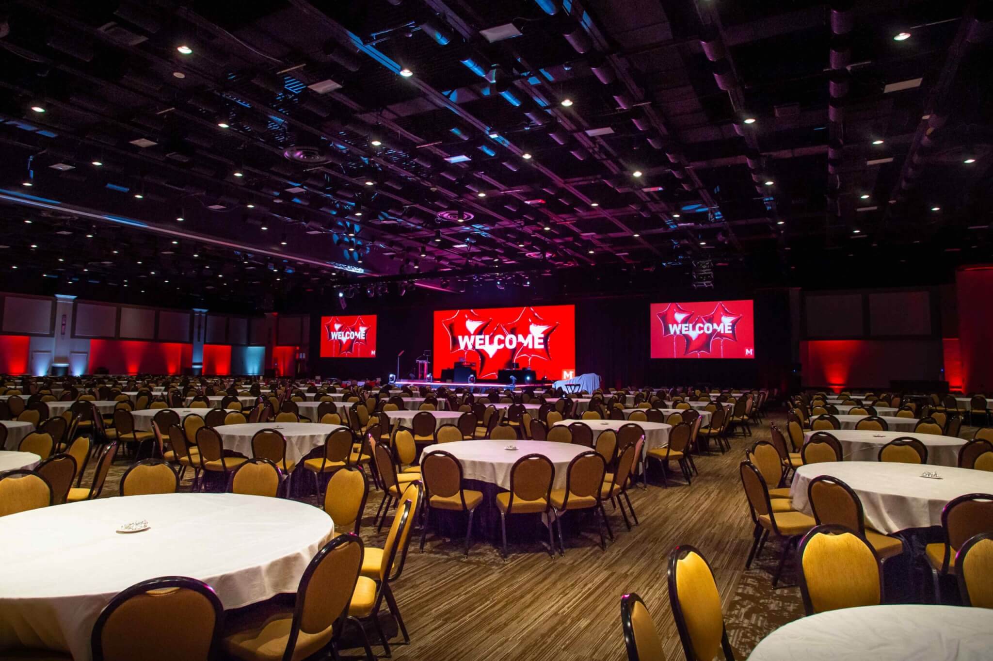 A large conference room with round tables and chairs set up, featuring red lighting and three screens displaying the word "WELCOME.