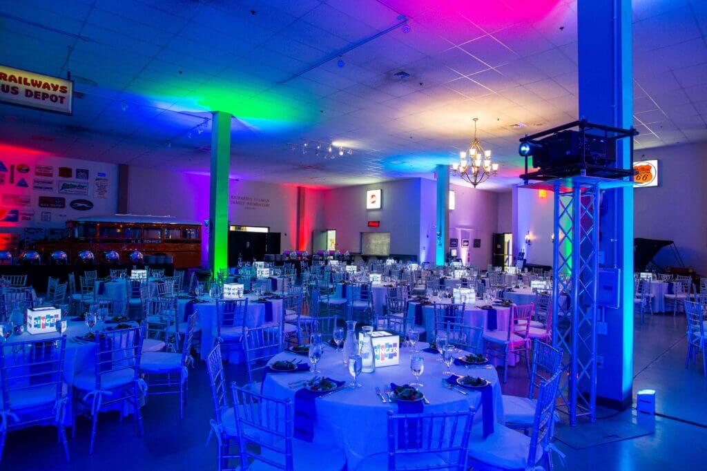 A large banquet hall with round tables set for an event, illuminated by colorful blue, pink, and green lighting.