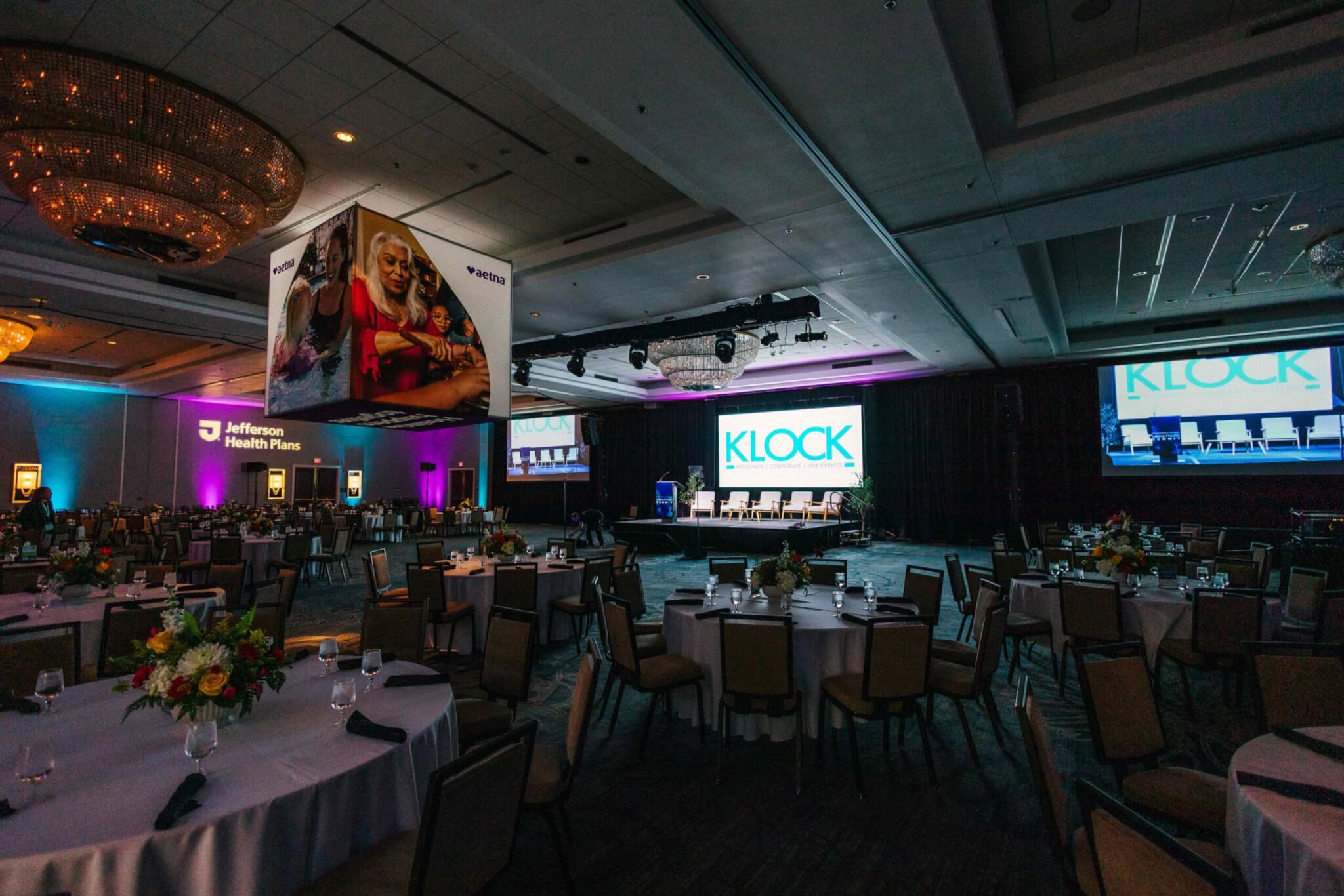 Adobe Express – file (3) A dimly lit banquet hall with round tables, floral centerpieces, and large screens displaying "KLOCK" and "Jefferson Health Plans" on stage.