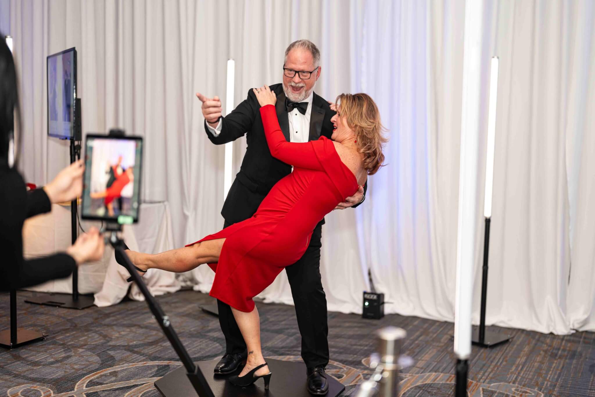 Adobe Express – file A man in a tuxedo dips a woman in a red dress while dancing at an event, with someone recording them on a smartphone.