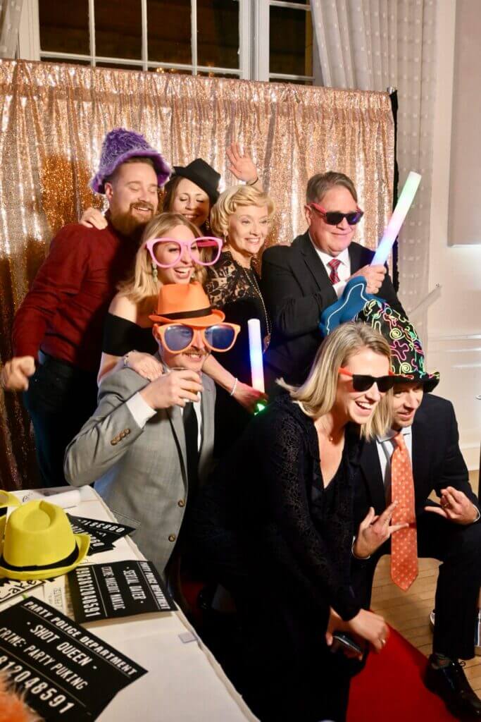 A group of adults pose together wearing colorful hats, oversized sunglasses, and holding glow sticks in front of a gold sequin backdrop at a party.