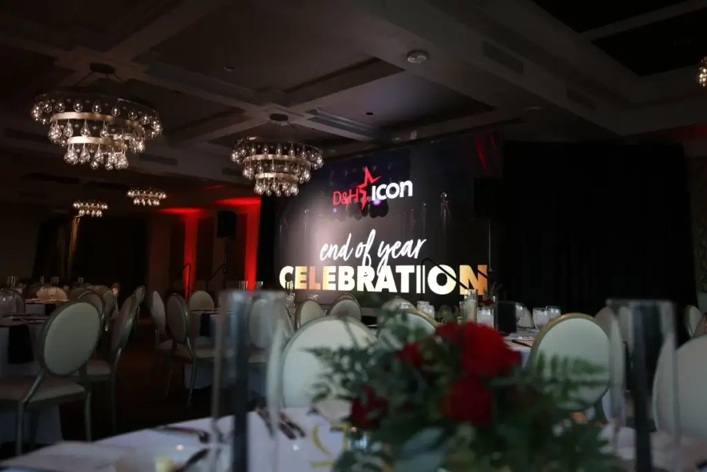 A decorated banquet hall with round tables and chairs set for an event; a stage in the background displays “end of year CELEBRATION” and “D&H Icon.”.