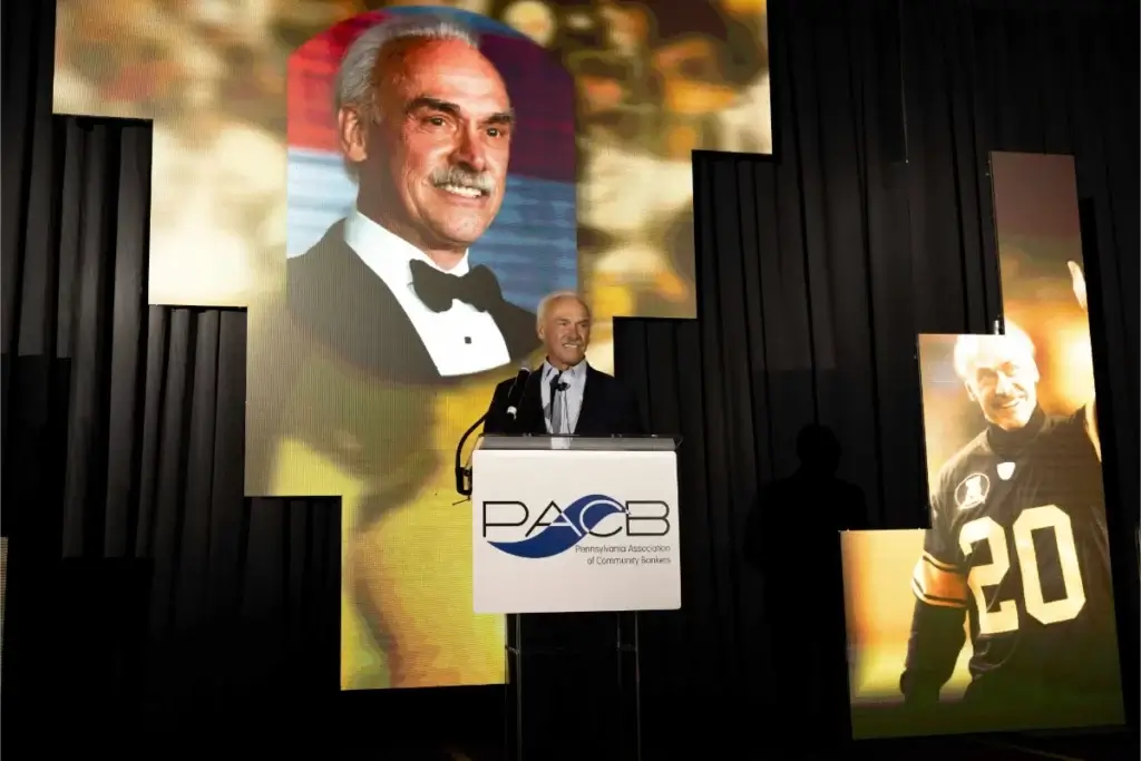 A man stands at a podium with a PACB sign, speaking at an event. Large images of him in a tuxedo and a football uniform are displayed in the background.