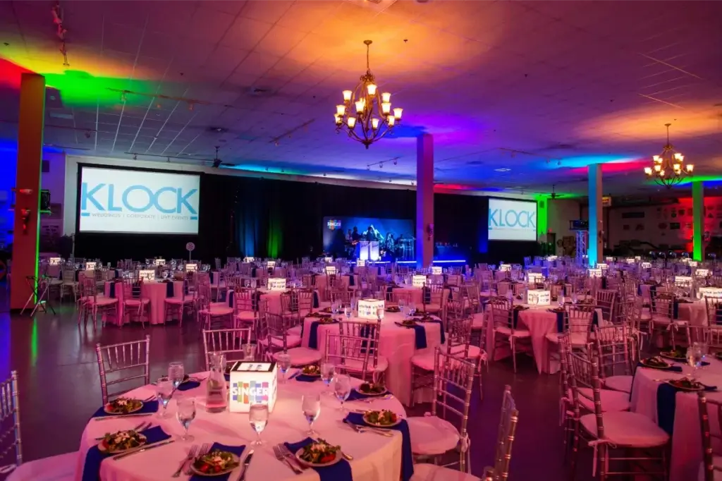 A large event hall set up for a formal dinner with round tables, clear chairs, colorful lighting, and projection screens displaying the Klock logo.