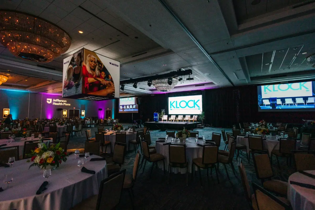 A large conference room set up with round tables, floral centerpieces, and a stage with screens displaying "KLOCK"; overhead banners and colorful lighting are visible.