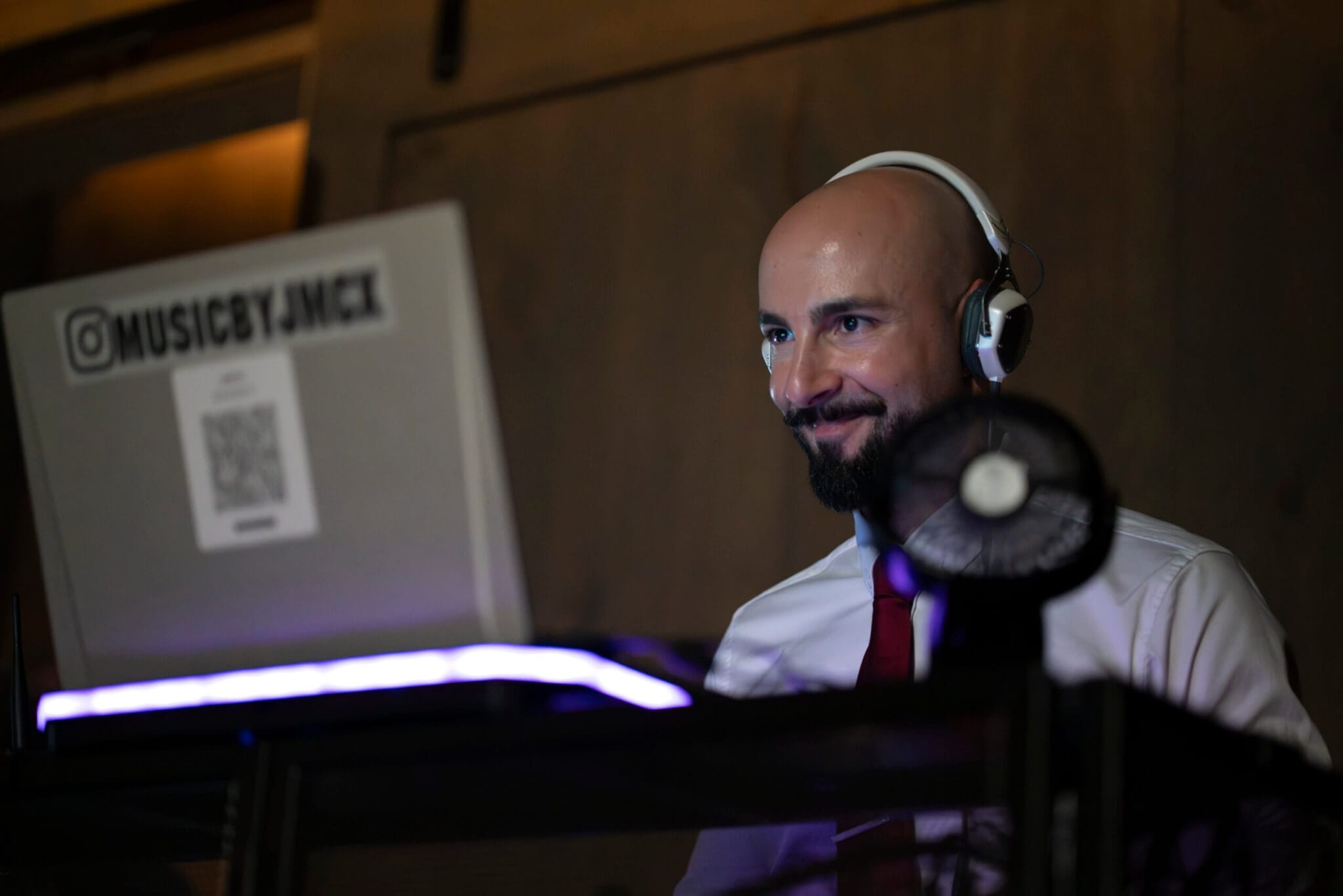 A man with headphones sits behind a laptop labeled "@MUSICBYJMCX," smiling, with a small fan and purple light on the desk in front of him.