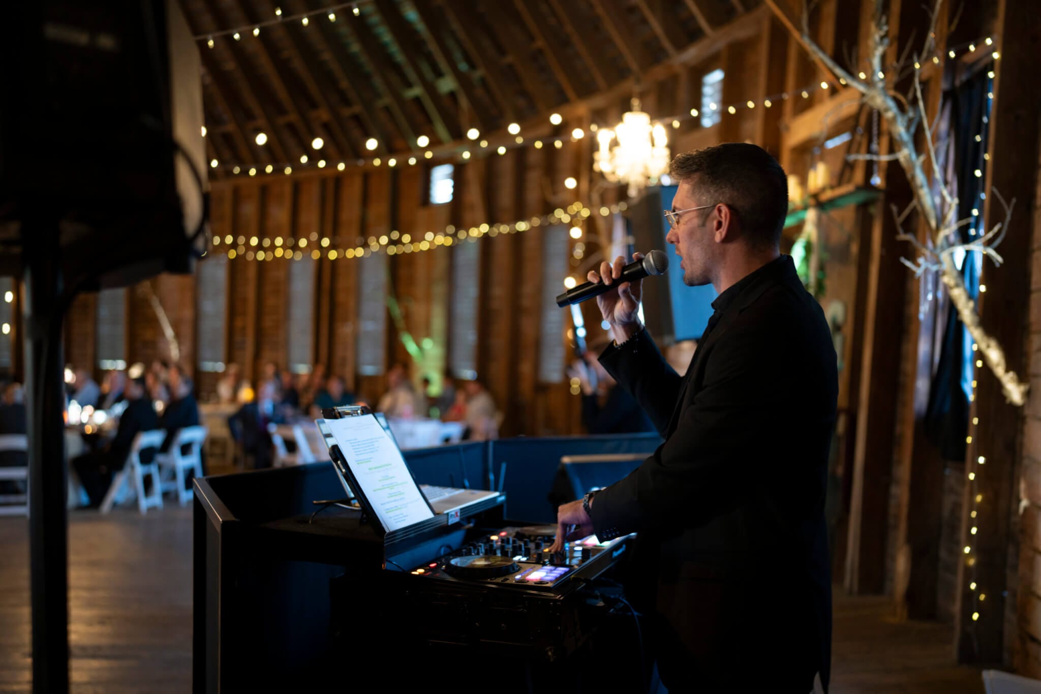 A DJ stands at a mixing console with a microphone, performing at an indoor event in a rustic venue decorated with string lights.