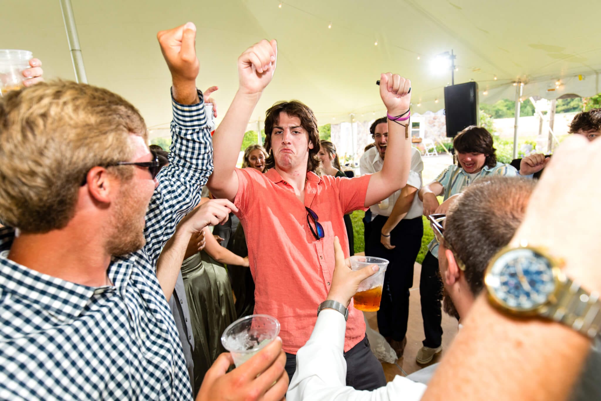 A man in a coral shirt dances enthusiastically with a group of people around him at an outdoor event under a large tent.