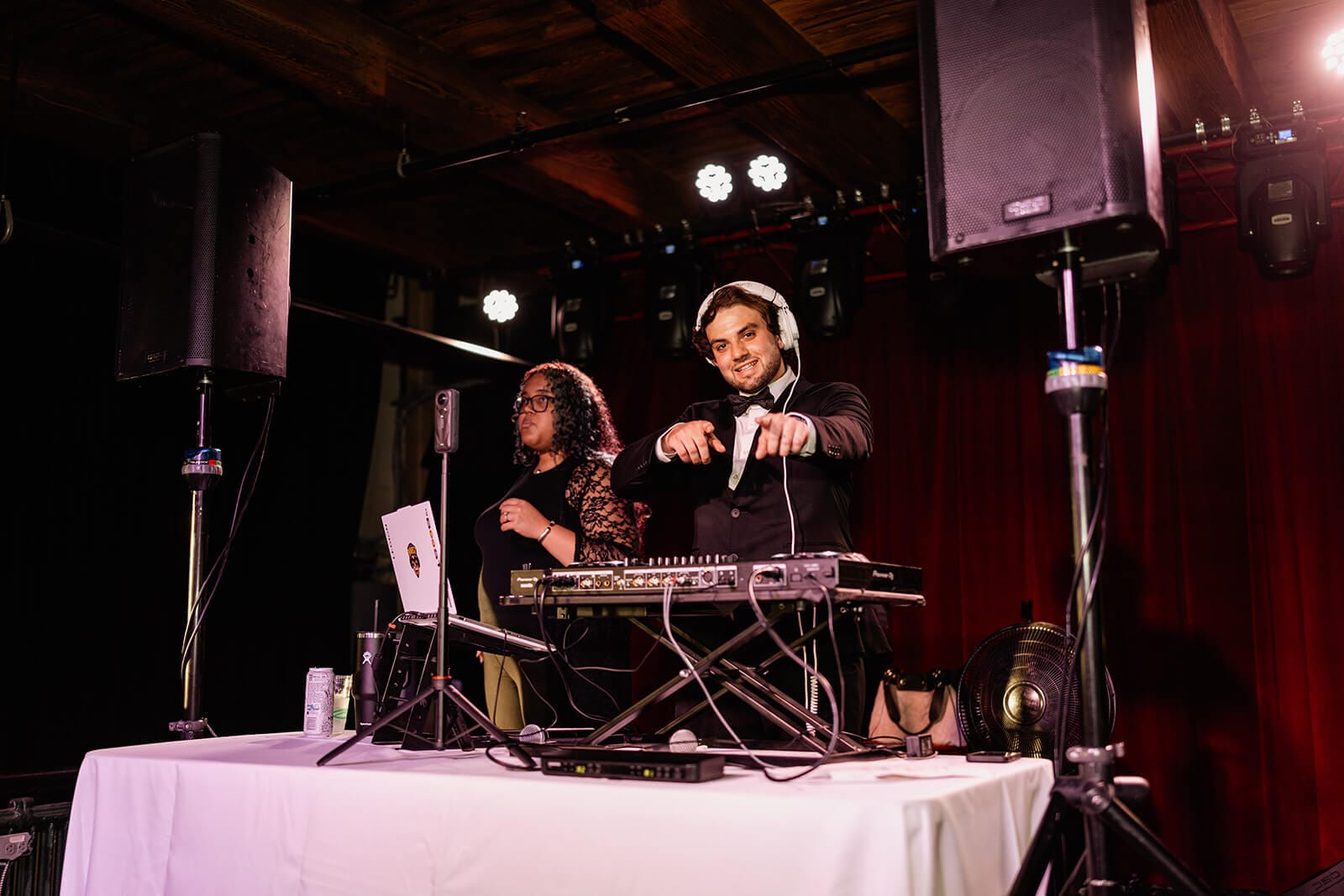 A man wearing headphones stands behind DJ equipment and points at the camera, while another person stands beside him. They are on a stage with red curtains and stage lights above.
