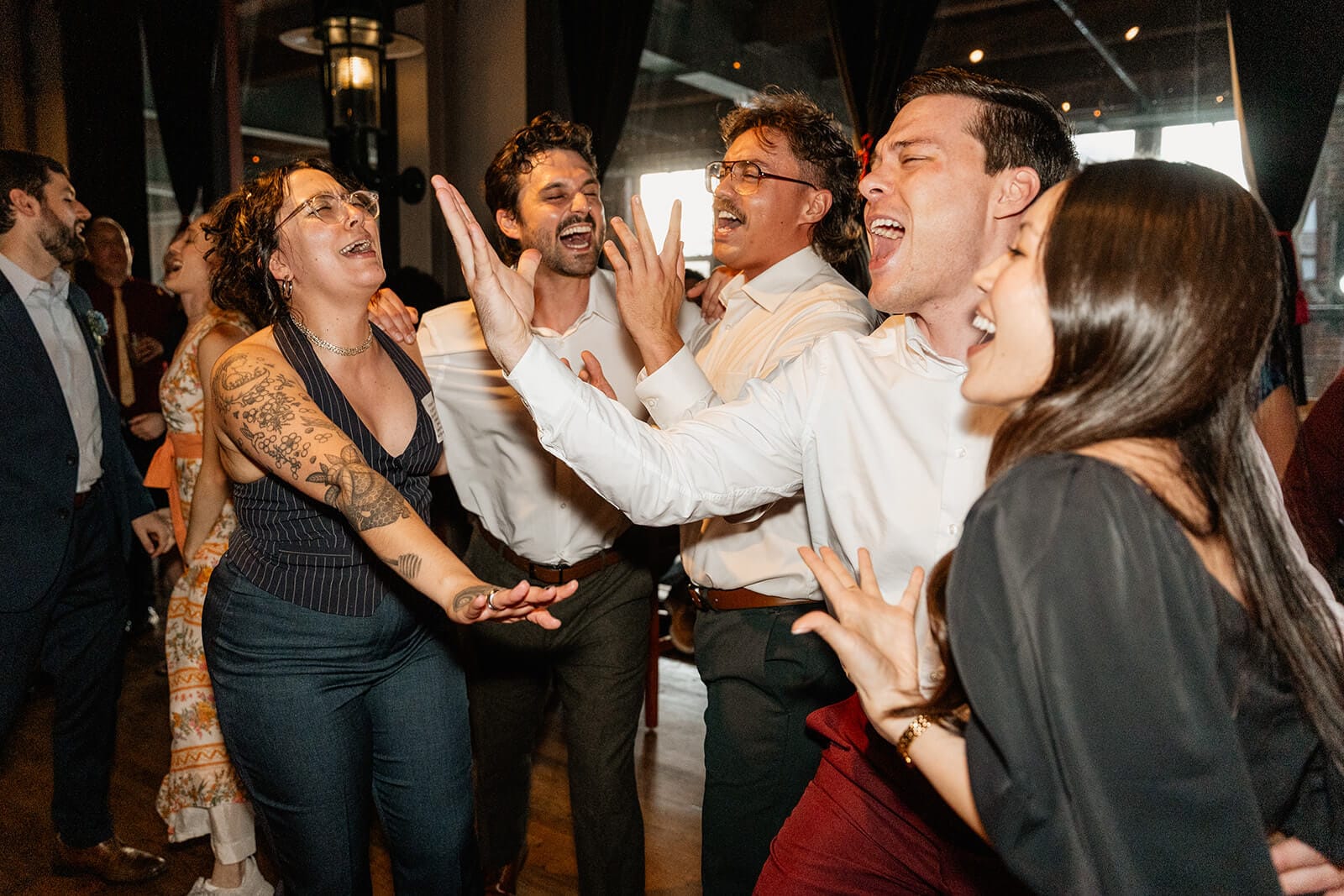 A group of people dressed in semi-formal attire enthusiastically sing and dance together at an indoor event or party.