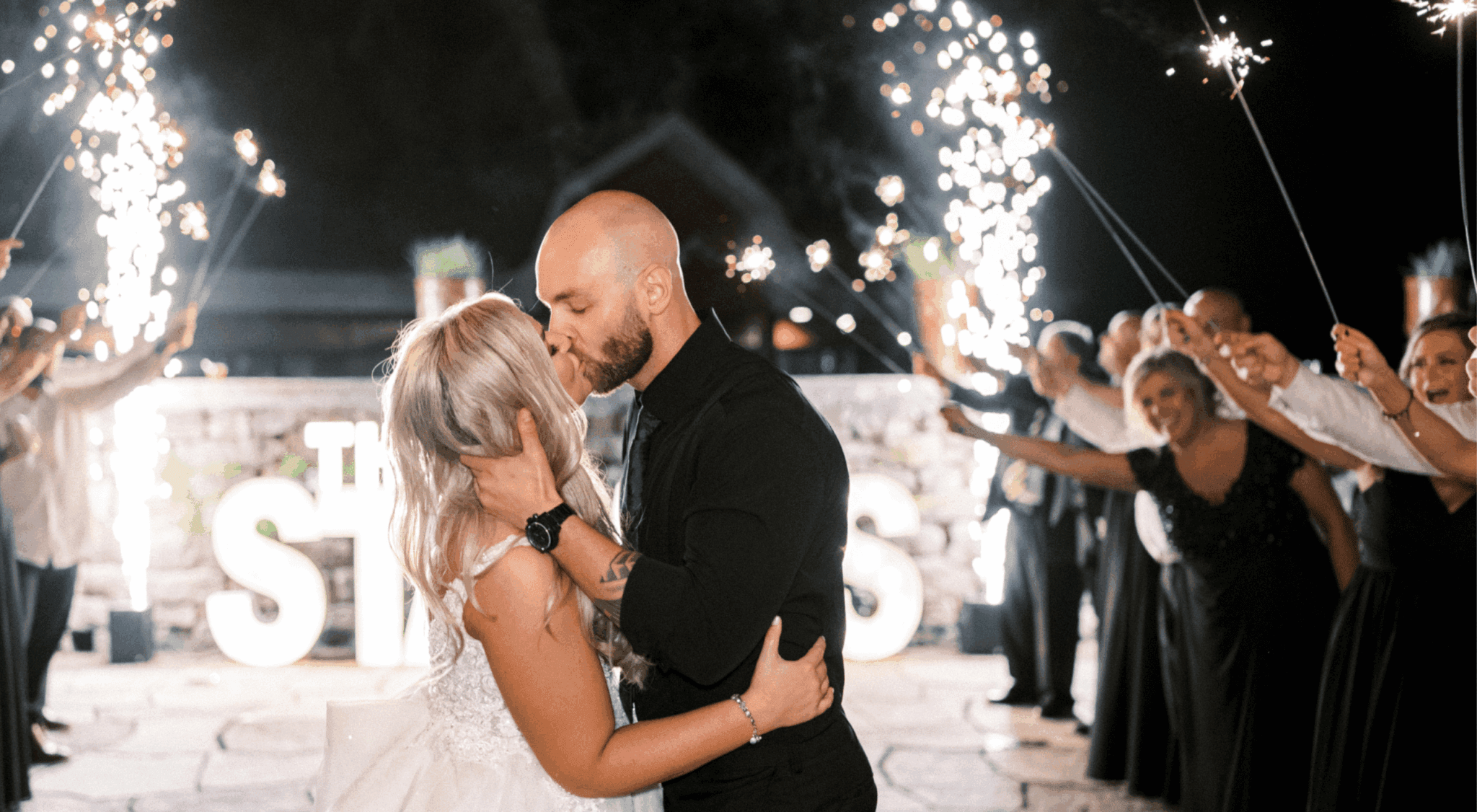 A couple kisses on a stone patio at night while guests hold sparklers and smile in celebration. Large illuminated letters are visible in the background.