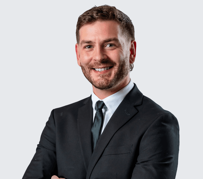 Man in a black suit and tie with arms crossed, smiling, facing forward against a plain light gray background.