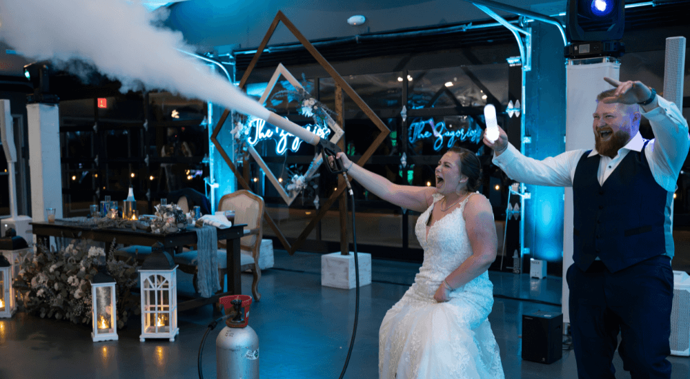 A bride, seated, uses a fog machine to spray smoke while a groom stands beside her cheering at a decorated indoor wedding reception.