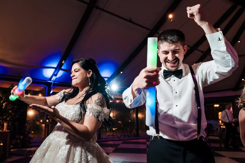 A bride and groom in formal attire dance energetically at a reception, each holding glowing LED foam sticks under colorful lighting.