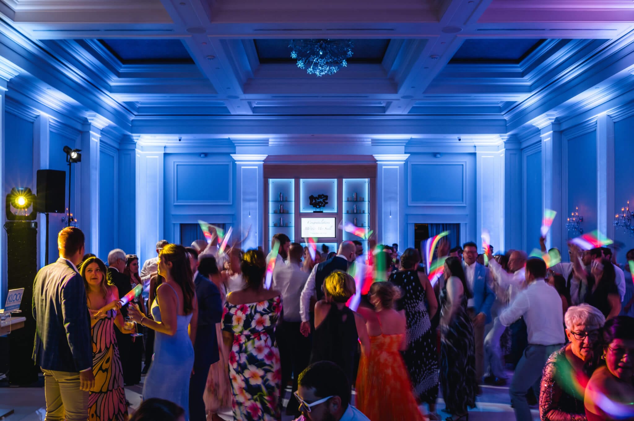 A large group of people dance and wave glow sticks in a spacious, well-lit ballroom with blue accent lighting.