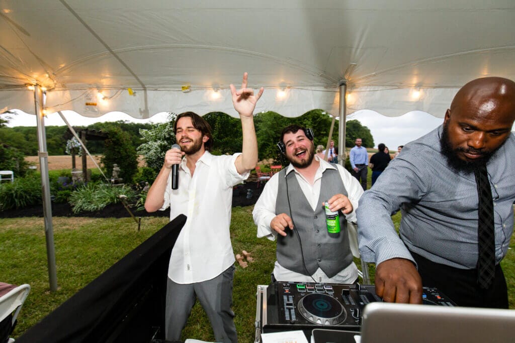 Three men stand under a white event tent. One holds a microphone with his hand raised, another smiles holding a can, and the third adjusts DJ equipment. People and greenery are in the background.