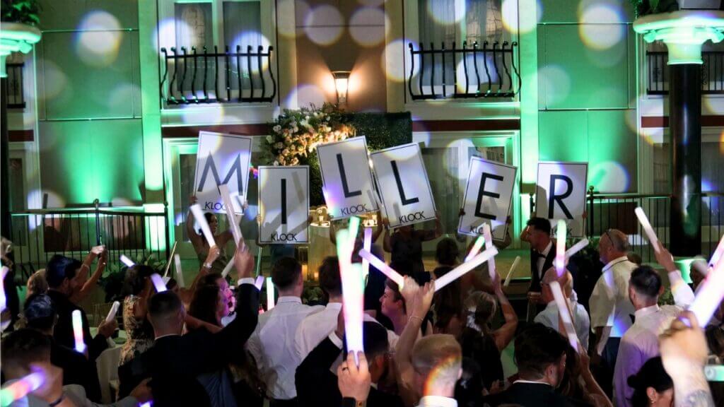 A crowd holds up signs spelling “MILLER” and waves light sticks at an indoor event with colorful lights and a decorated backdrop.