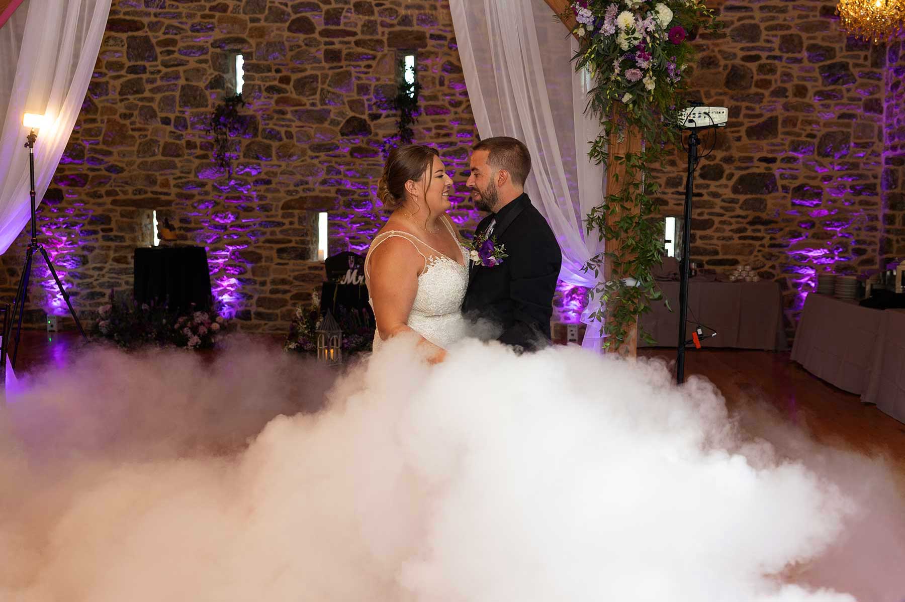 A bride and groom share a dance surrounded by floor fog, with a stone wall and purple lighting in the background.