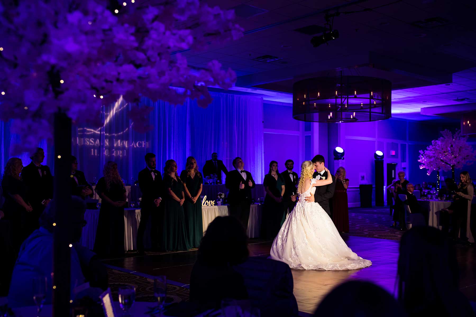 A bride and groom share their first dance on a dimly lit ballroom floor, surrounded by their wedding party and guests under purple lighting.