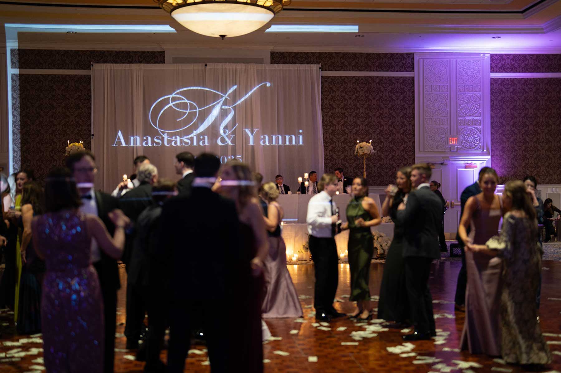 People in formal attire dancing at a wedding reception with "Anastasia & Yanni" projected on a backdrop in a decorated ballroom.