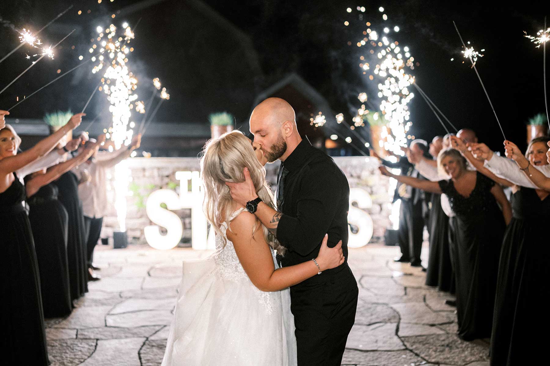 A bride and groom share a kiss outdoors at night, surrounded by guests holding sparklers, with a large illuminated sign in the background.