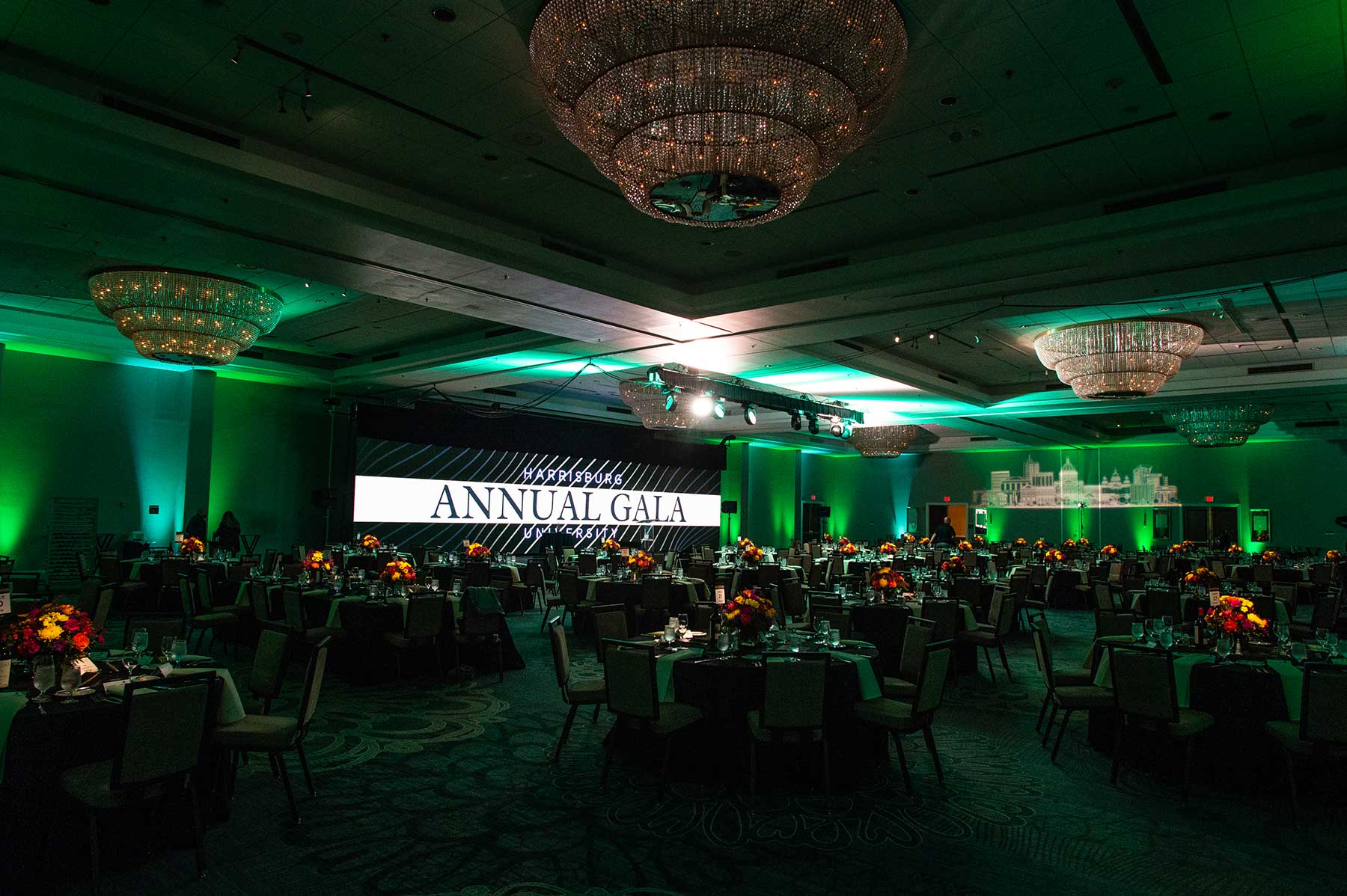 corporate event annual gala dj A large ballroom set up for an event, with round tables, floral centerpieces, chandeliers, and a stage displaying "Annual Gala" on a large screen.