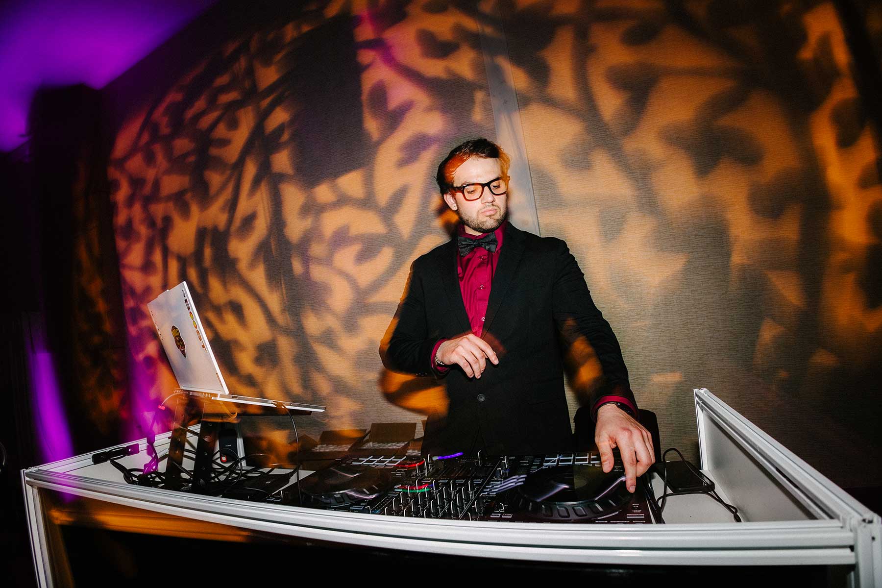 dj at event with pattern projection lighting A man in a suit and glasses stands behind a DJ booth, adjusting equipment, with patterned lighting on the wall in the background.