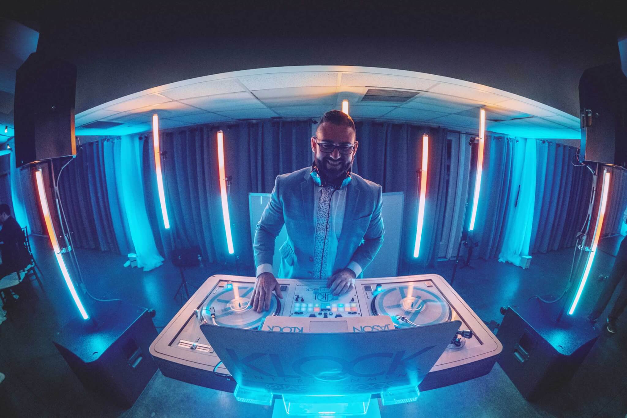 A DJ in a suit stands behind a booth with turntables, performing in a room with blue lighting and vertical LED light bars.