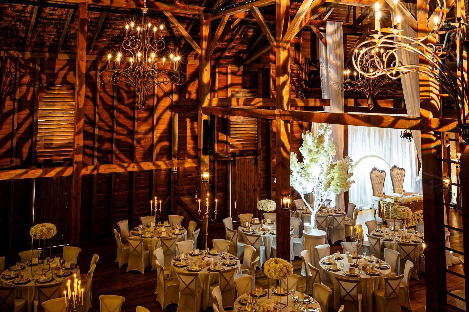 Elegant indoor wedding reception in a rustic barn with round tables, white tablecloths, floral centerpieces, chandeliers, and decorative lighting casting patterns on wooden walls.