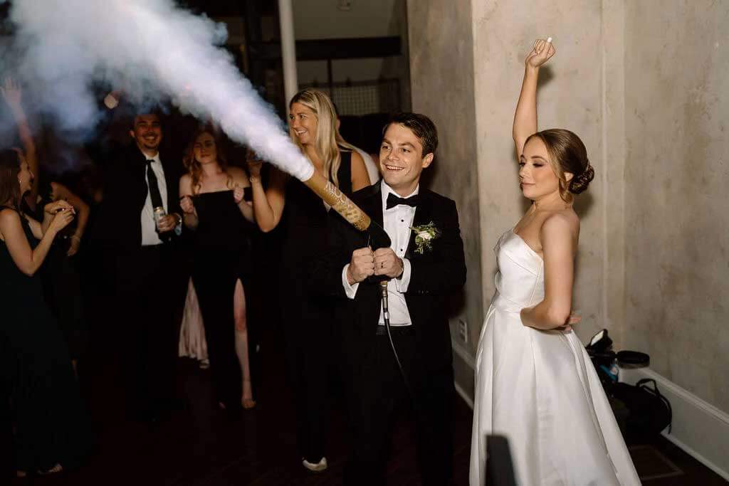testimonial-caitlyn A groom in a tuxedo uses a CO2 cannon, emitting smoke, while the bride in a white dress stands beside him with her arm raised; guests watch in the background.
