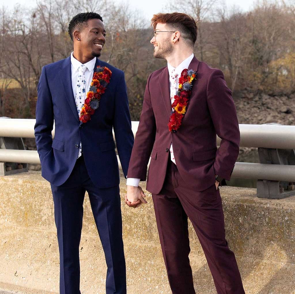 testimonial-dorrin-alt Two men in suits stand on a bridge holding hands, each wearing a boutonniere with red and orange flowers, smiling at each other.