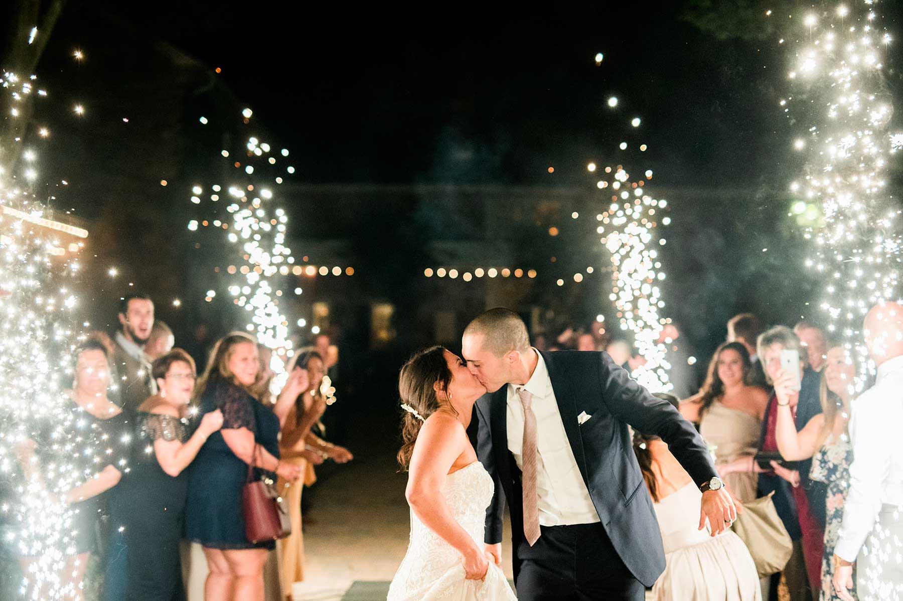 A bride and groom kiss while walking through a pathway lined with guests and sparklers at night.
