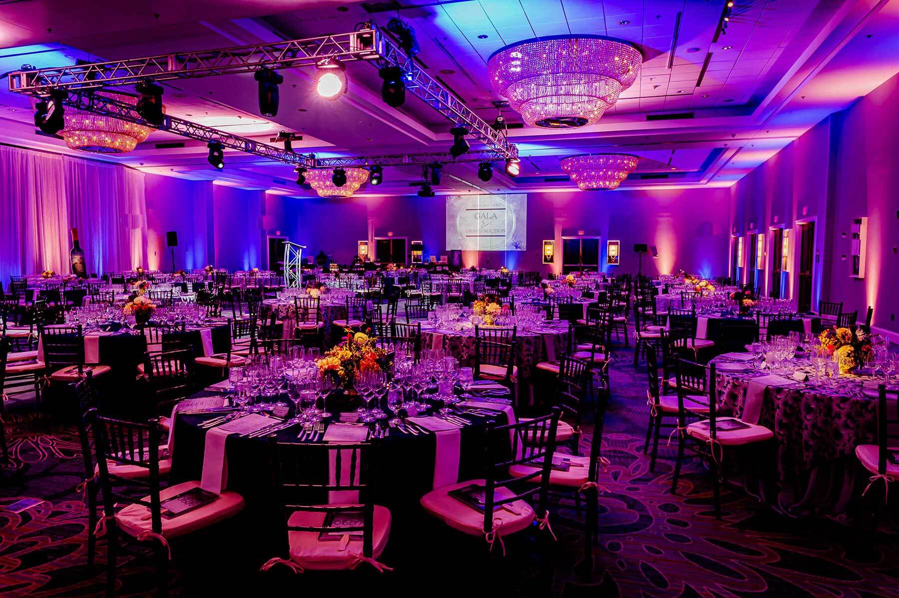 A large banquet hall set for an event with round tables, floral centerpieces, purple lighting, and projection screens at the front.