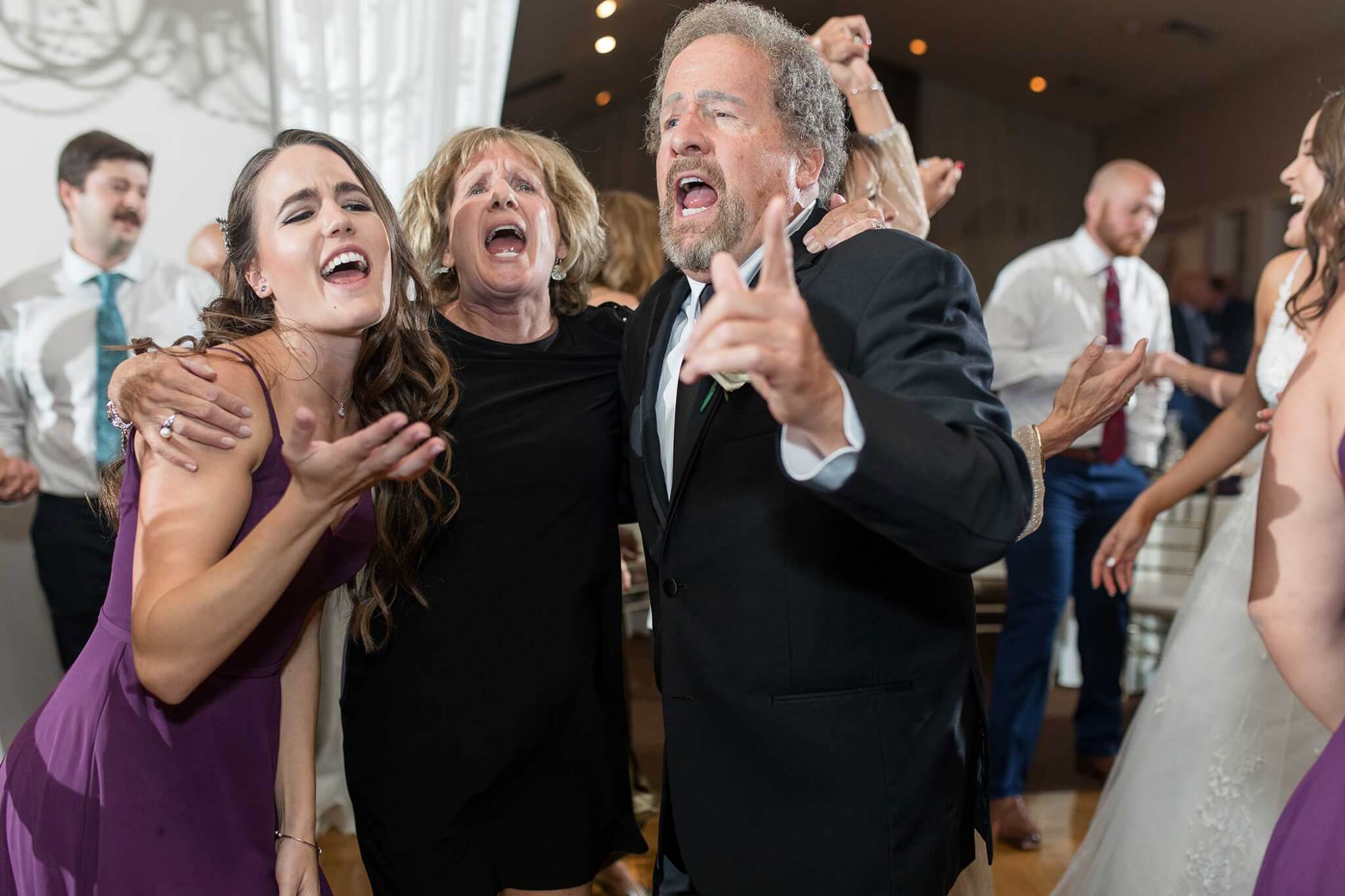 wedding-guests-on-dance-floor-at-The-Links-at-Gettysburg A group of people, including a man in a suit and two women in dresses, sing and celebrate together at an indoor event or party.
