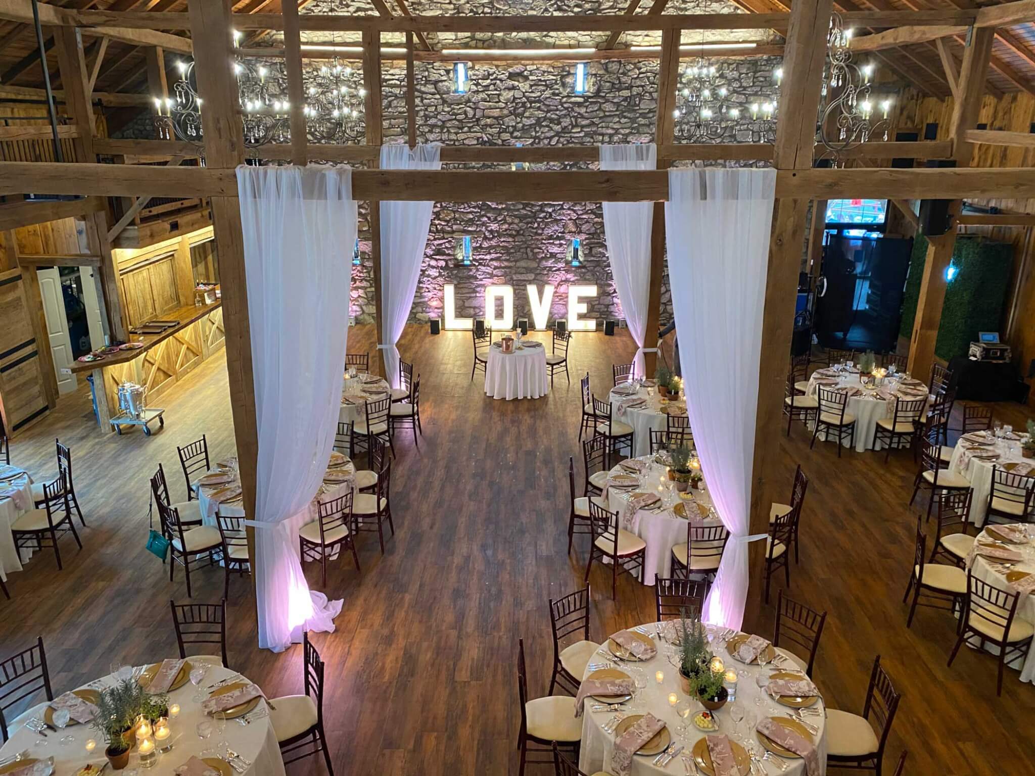 A decorated barn venue with round tables set for an event, white drapes, and large illuminated "LOVE" letters against a stone wall at the back.