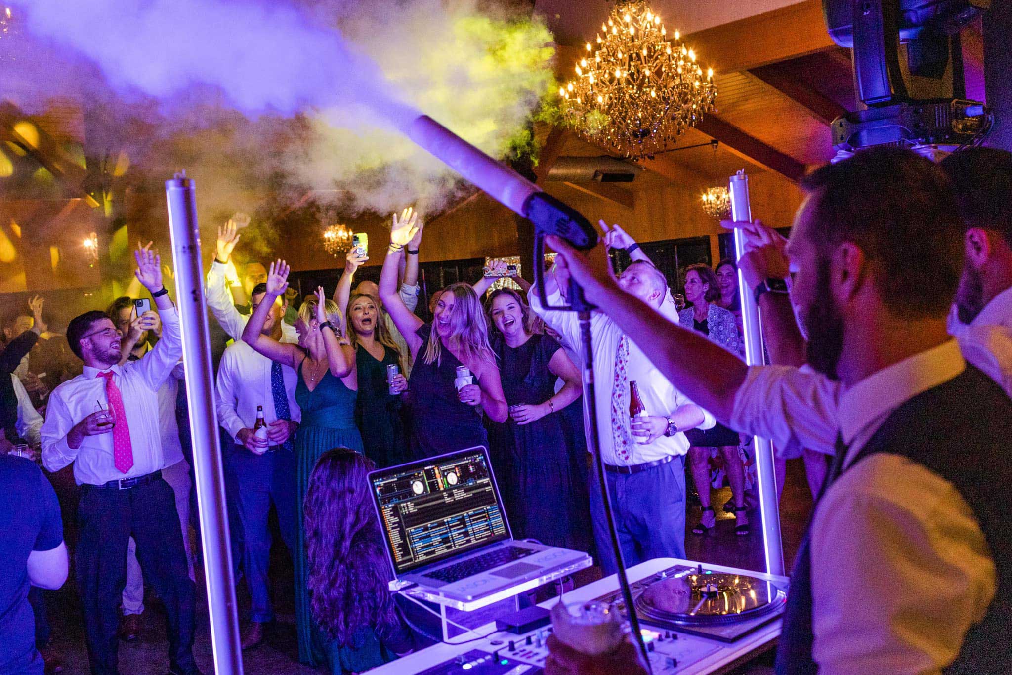 CO2-cannon-Historic-Acres A DJ plays music while a crowd of people in formal attire dance and raise their hands at an indoor party with bright lights and fog effects.