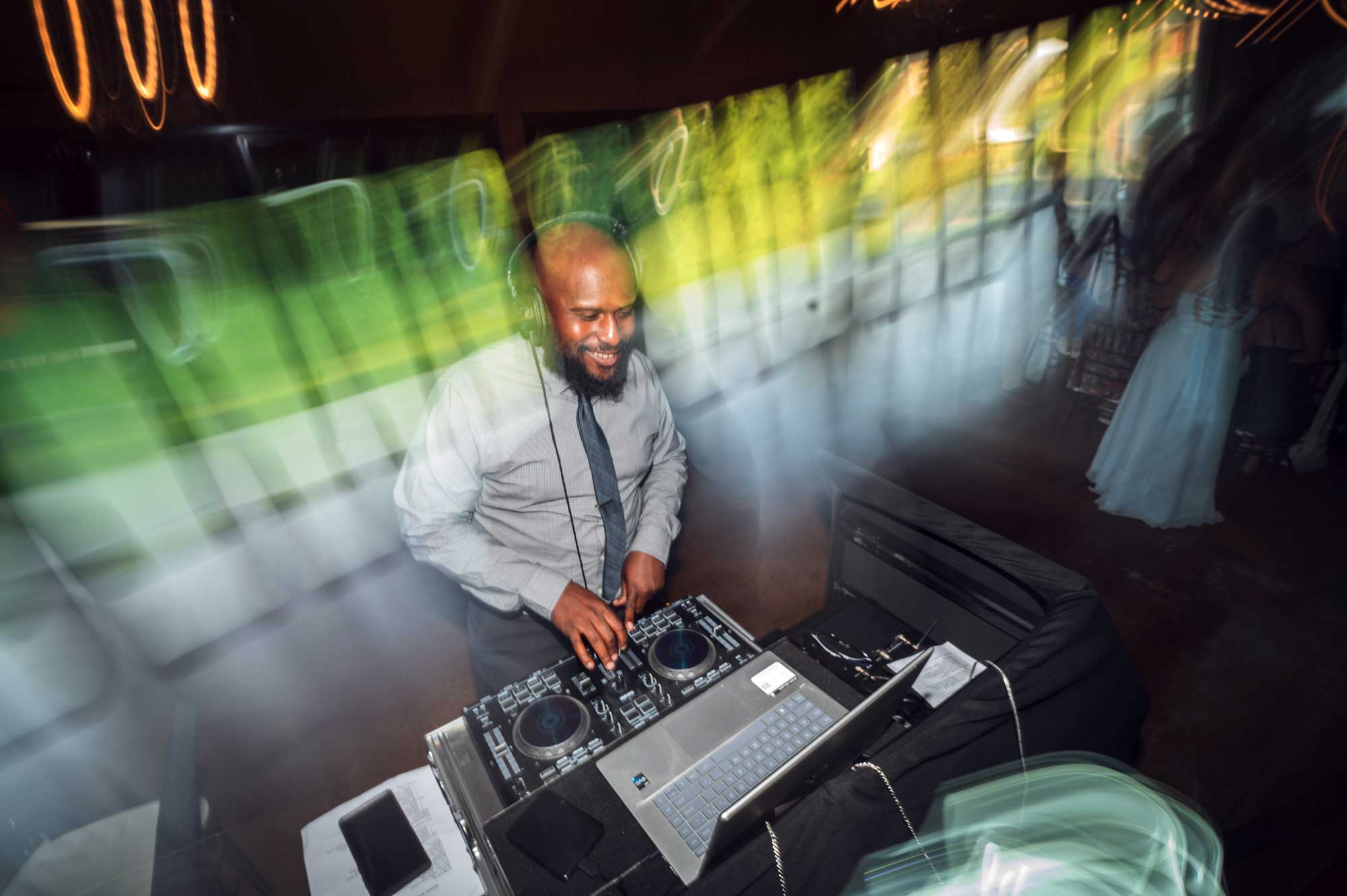 Historic-Acres-Klock-DJ-Dephilip-Jackson A DJ in a dress shirt and tie mixes music on a DJ controller, surrounded by colorful motion blur and a laptop at an indoor event.