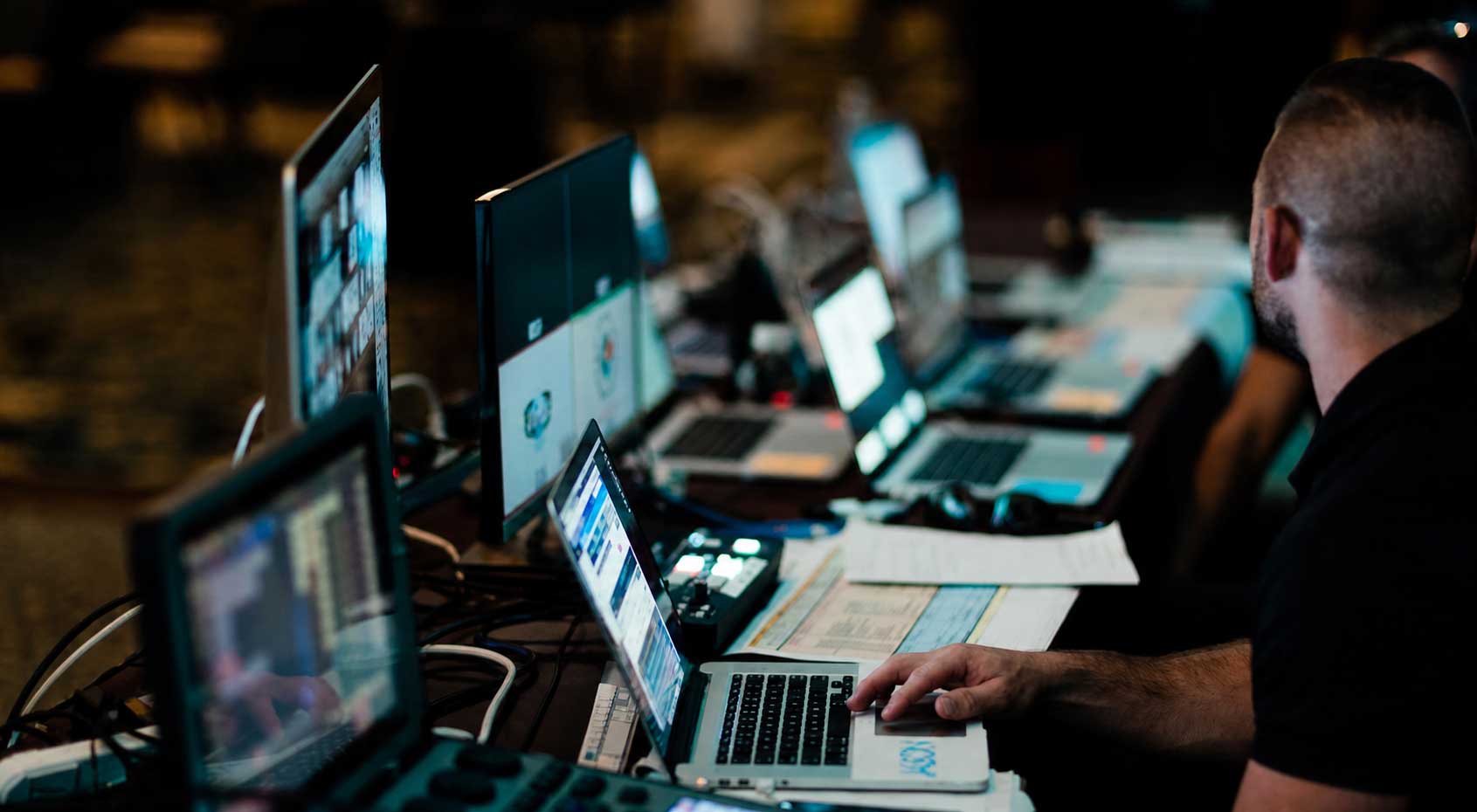 Klock-at-Work Person sitting at a desk with multiple laptops, monitors, and electronic equipment, appearing to manage or monitor various digital tasks.