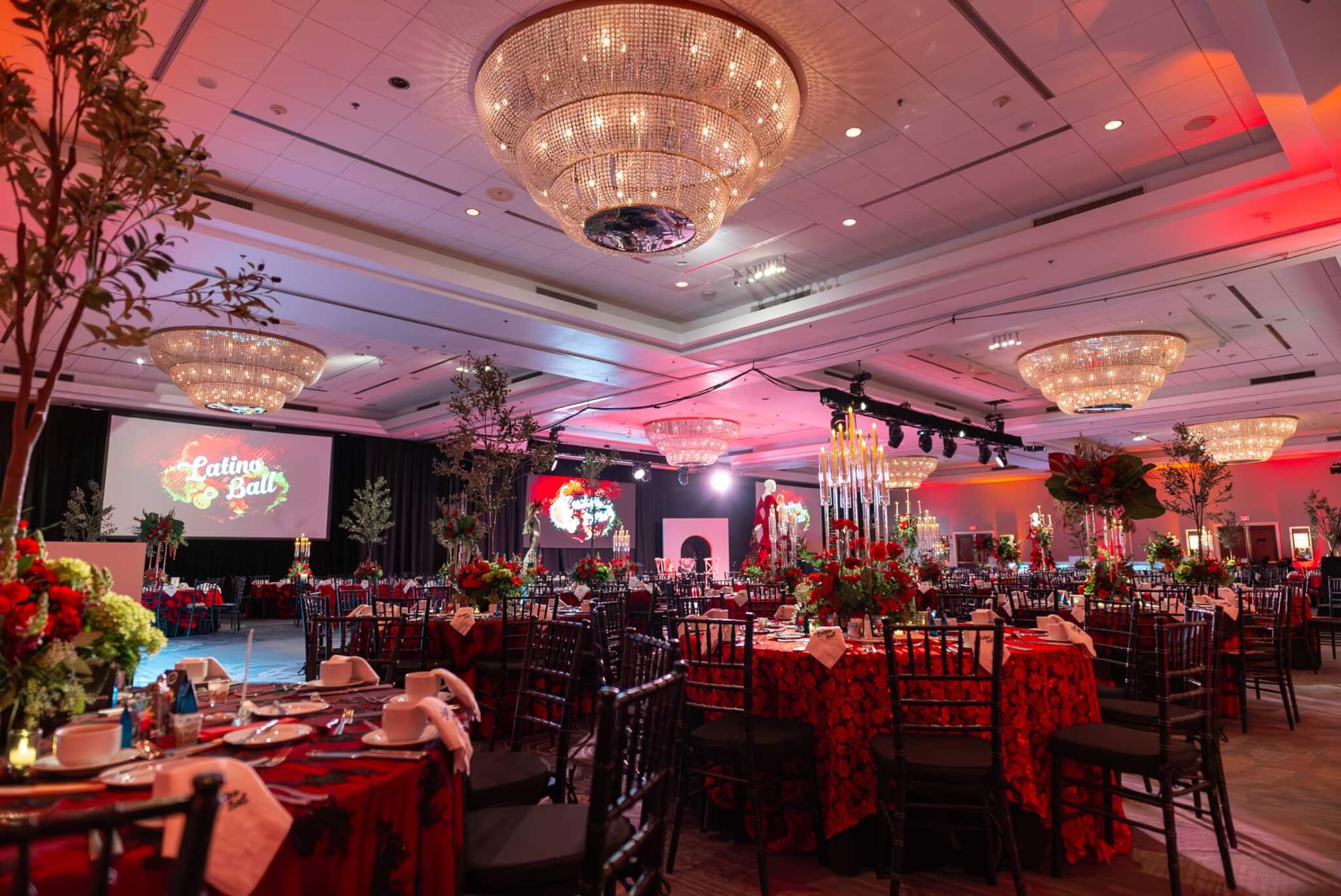 Latino-Ball-Hilton-Harrisburg-KlockEntertainment_ Ballroom set for an event with round tables covered in red floral tablecloths, elaborate centerpieces, chandeliers, and a large screen displaying “Latino Ball.”.