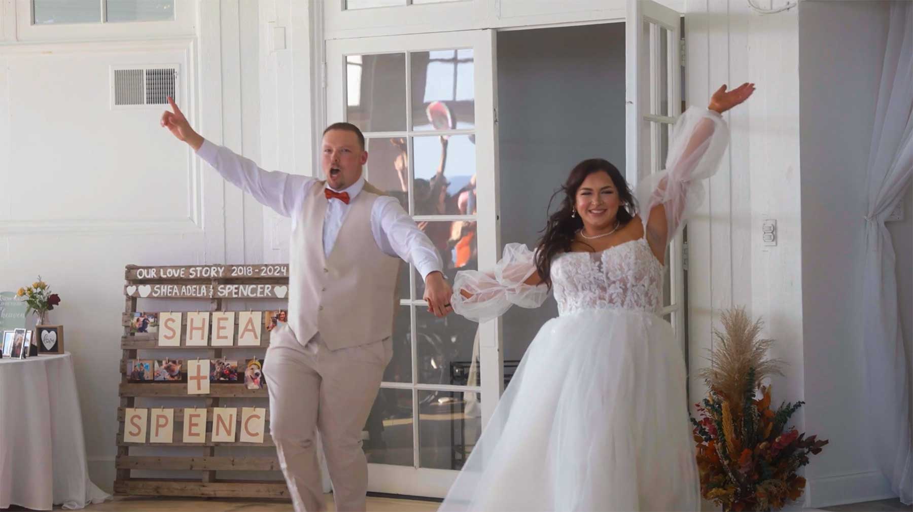 Rotunda-at-Lauxmont-Farms-wedding-couple-entrance A bride and groom enter a bright room holding hands, both smiling with arms raised. A decorated wooden sign and floral arrangement are visible in the background.