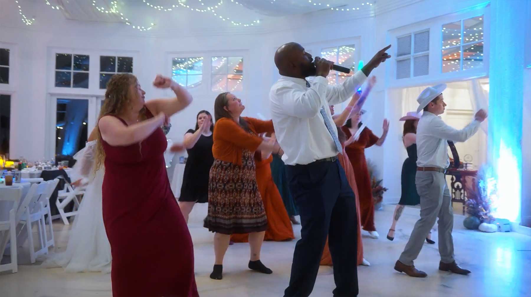 Rotunda-at-Lauxmont-Farms-wedding-dj-dance A group of people dance in a decorated indoor venue, following a man in a white shirt and tie who is holding a microphone and leading the group.